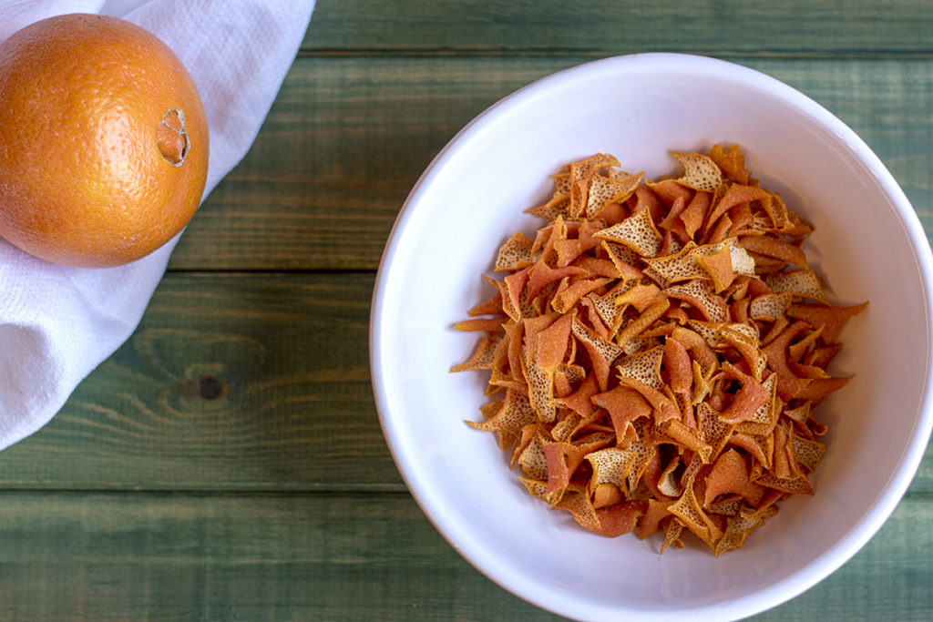 A bowl of dried orange peels and an orange on a white tea towel rest on a green tabletop.