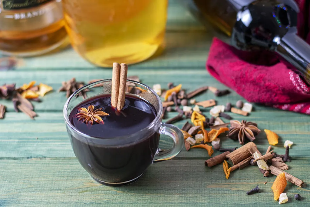 A mug of hot mulled wine next to mulling spices scattered on a green wood tabletop. A bottle of wine, honey, and brandy are in the background.