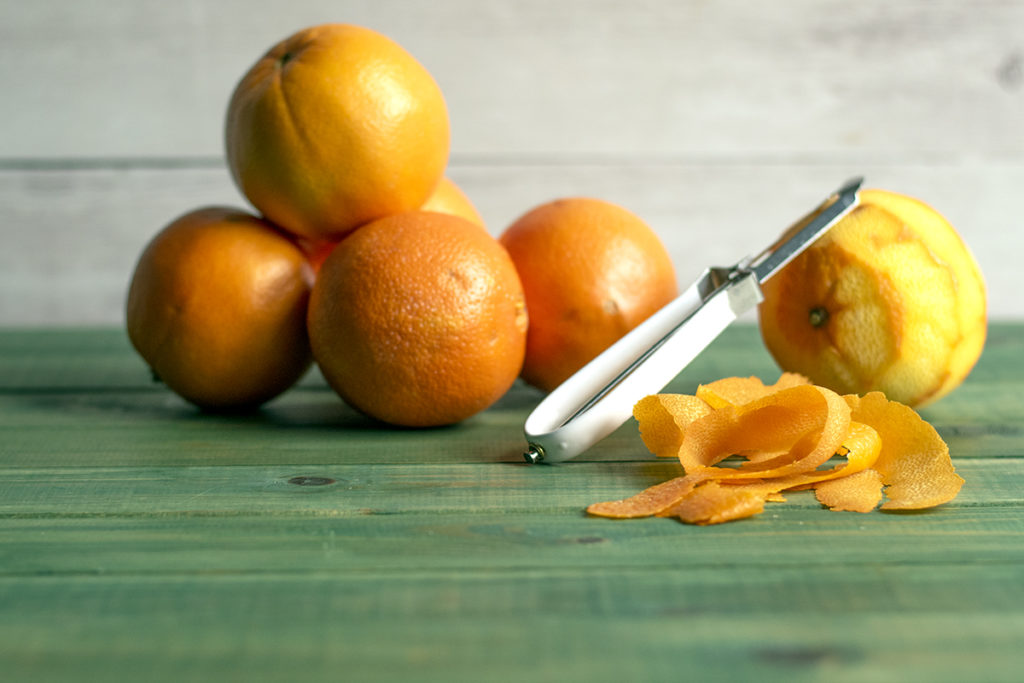 A bunch of oranges are stacked on a green tabletop. One orange, which has been peeled, has a vegetable peeler resting on it. 