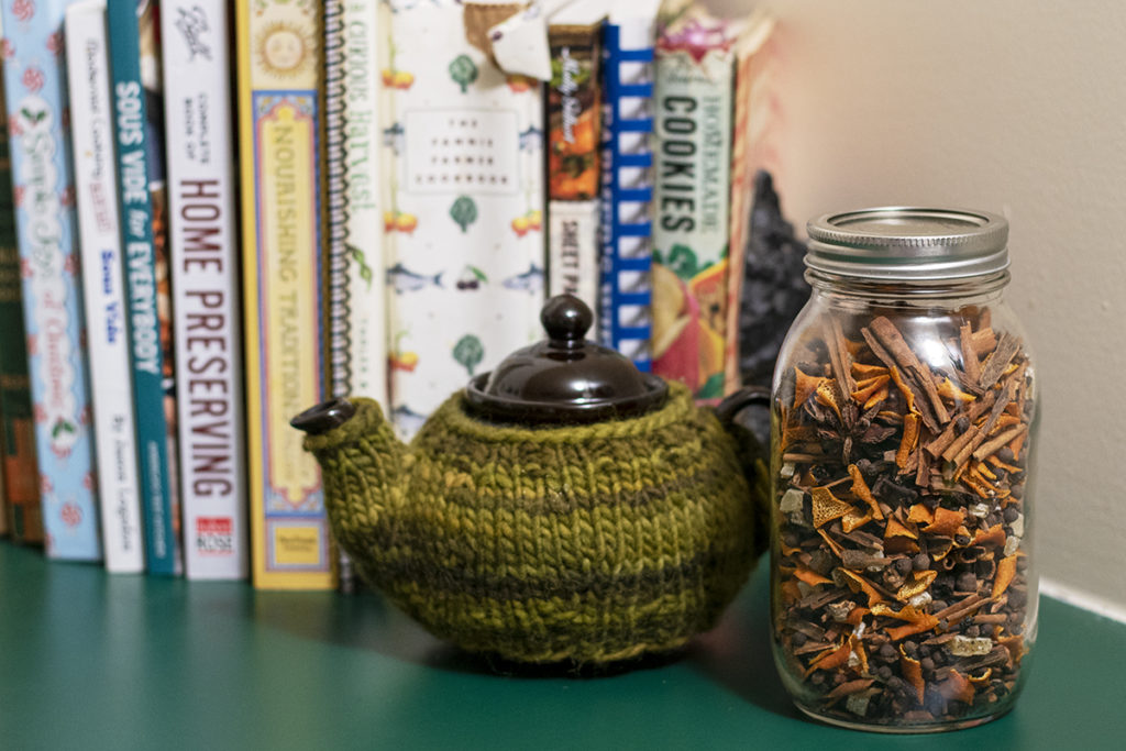 A jar of mulling spices is next to a tea pot on a green kitchen counter. There are cookbooks in the background.