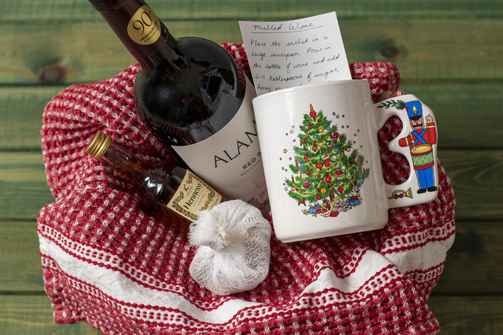 A gift basket with a red kitchen towel. The basket contains a bottle of wine, a tiny bottle of cognac, a sachet filled with mulling spices and Christmas mug mug with a notecard tucked inside. The mug has a Christmas tree on it and the handle has a toy soldier.