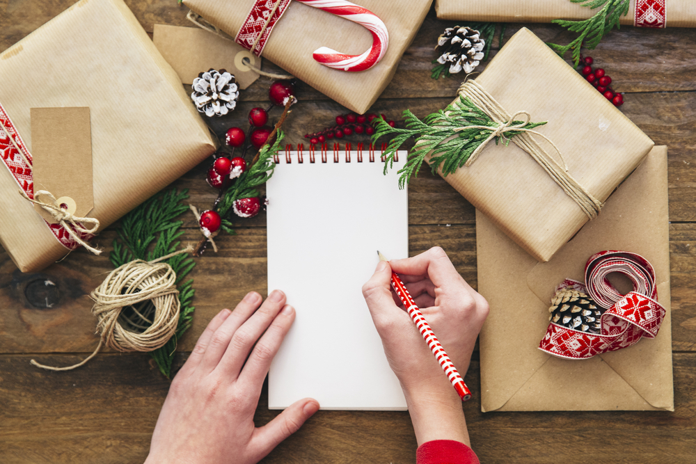 Persons hands shown writing letter on blank notepad, wrapped Christmas presents nearby.