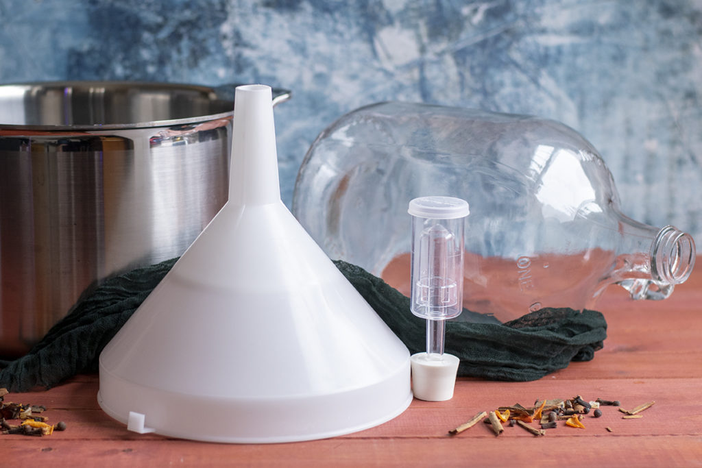 A stockpot, a funnel with a strainer, a one gallon glass jug and a three piece airlock arranged on a wooden top. 