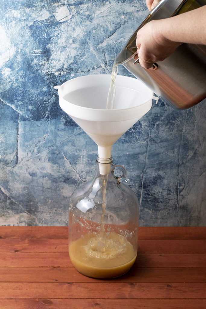 Someone pours the honey and water mixture from a stockpot into a funnel fitted into the glass jug. 