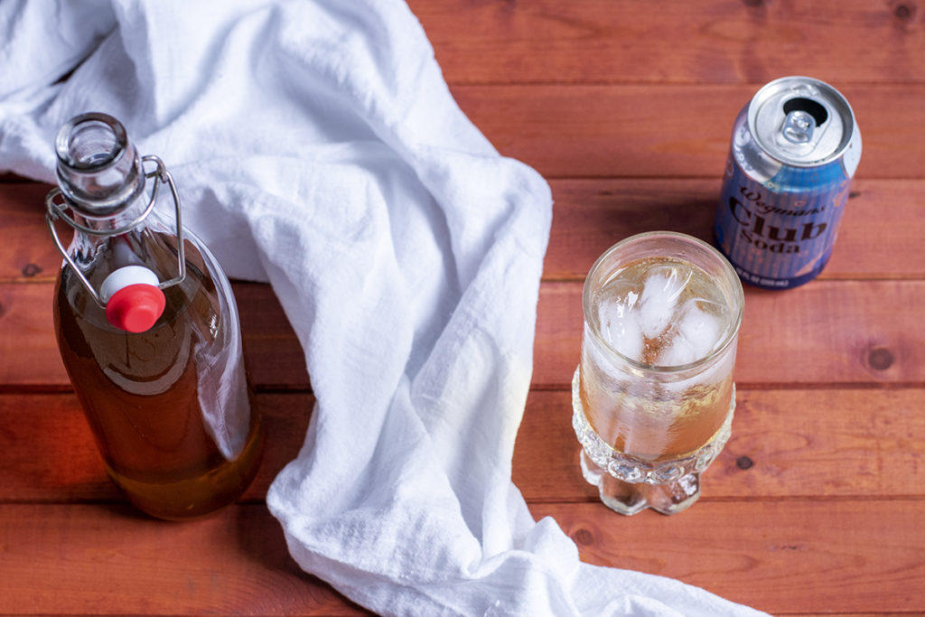A glass of club soda and fire cider on a wooden tabletop. There is a white kitchen towel and a can of club soda and a bottle of fire cider. 