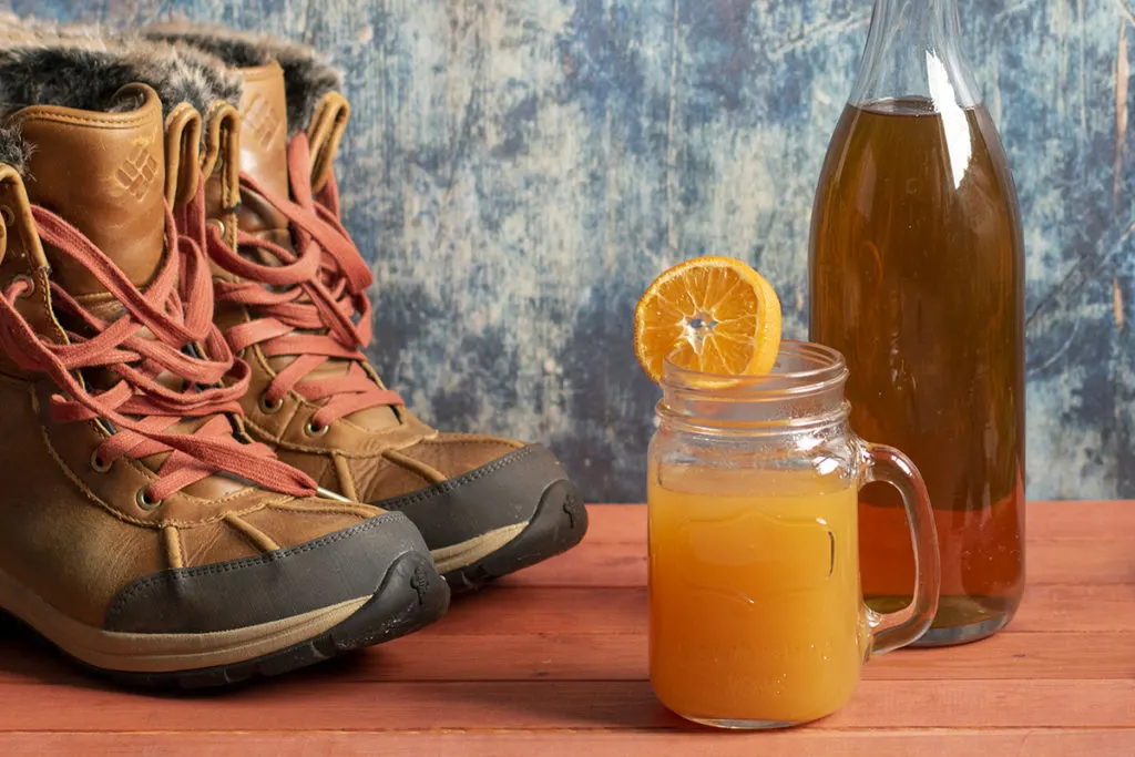 A glass mug of hot apple cider garnished with an orange slice, a bottle of fire cider next to it. To the side is a pair of snow boots. 