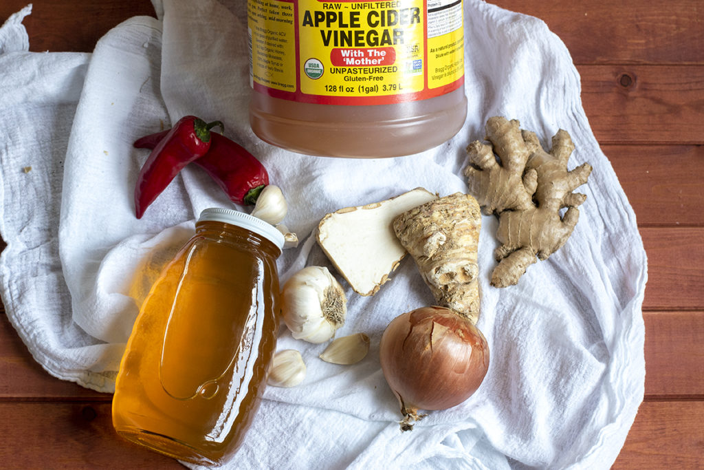 A large jug of apple cider vinegar, a jar of honey, hot peppers, garlic cloves, horse radish root, an onion and fresh ginger root are decoratively arranged on a white kitchen towel. 