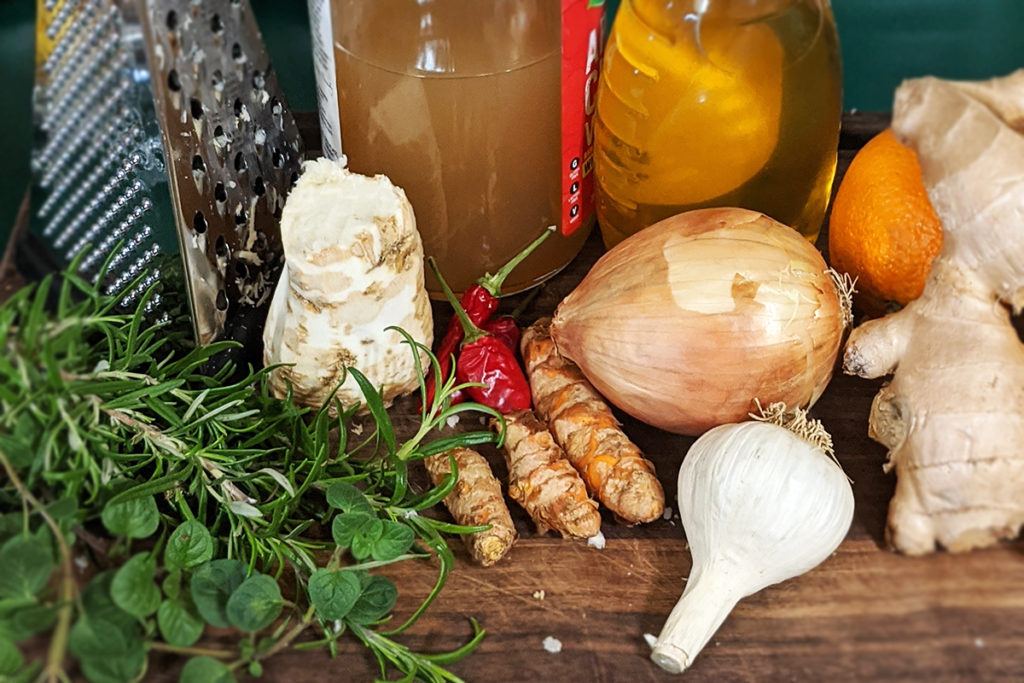 A cutting board with a cheese grater, a bottle of apple cider vinegar, and a jar of honey. There is also an orange, fresh ginger root, an onion, a bulb of garlic, fresh turmeric root, a couple of hot peppers, a chunk of horse radish rook and a few sprigs of rosemary and oregano.