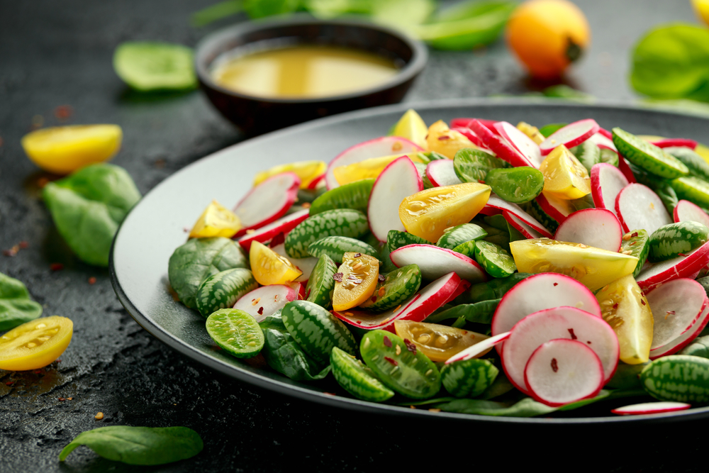 A fresh salad made out of sliced radishes, pear tomatoes, and cucamelons arranged on a black plate. 