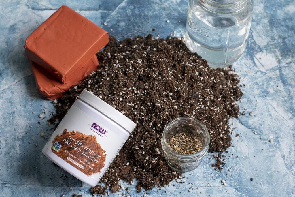 A pile of potting soil with a jar of water, a smaller jar of seeds, a tub of red clay powder and terracotta modeling clay surrounding it.
