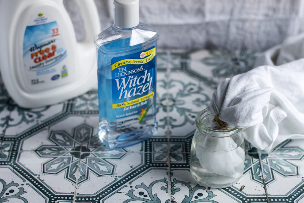 A portion of a t-shirt is soaking in a small jar of witch hazel on a tiled floor. There is a bottle of witch hazel and a bottle of laundry detergent next to the shirt.