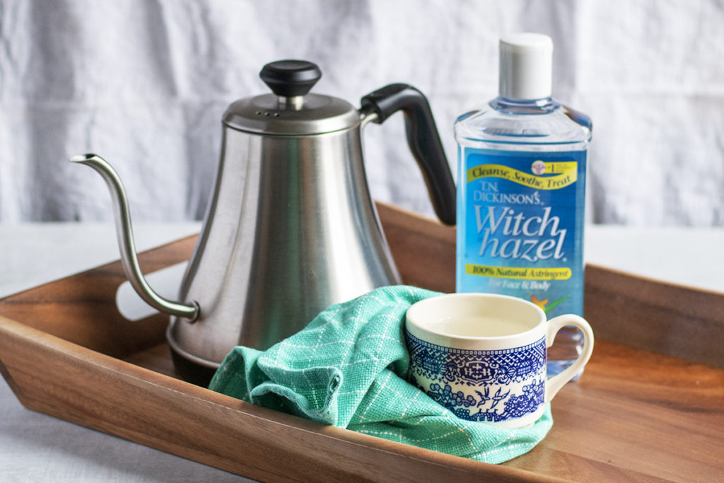 A tea kettle, a bottle of witch hazel, a tea towel and a tea cup are set on a tea tray.