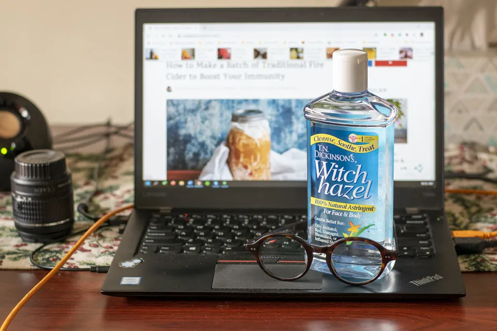 A desk with a laptop and a camera lens. A bottle of witch hazel and a pair of eyeglasses sit on the laptop. 