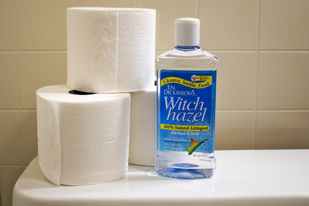 A bottle of witch hazel is sitting next to three rolls of toilet paper on the back of a toilet.