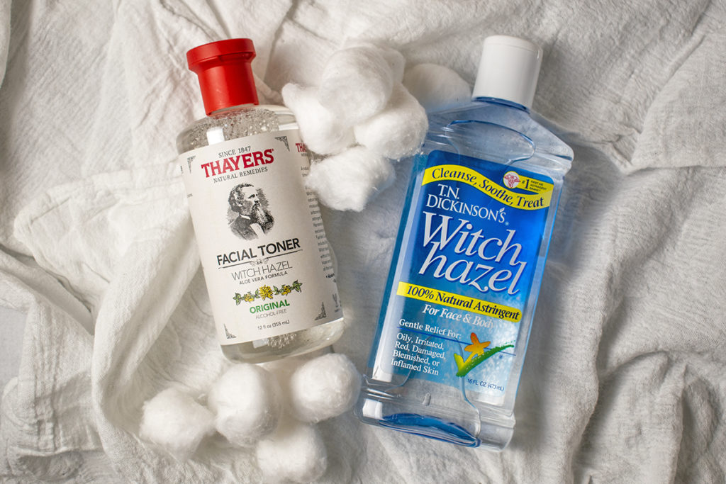 A bottle of Thayer's witch hazel and a bottle of T.N. Dickinson's Witch Hazel lay next to each other on a cloth background. There are two small piles of cotton balls.