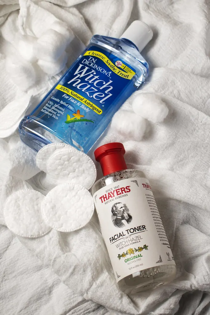 A bottle of Thayer's witch hazel and a bottle of T.N. Dickinson's Witch Hazel lay next to each other on a cloth background. There are two small piles of cotton balls.