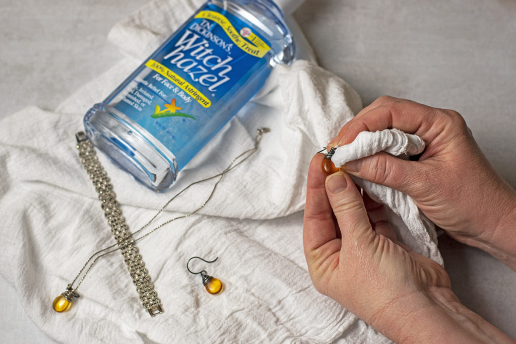 Hands are cleaning an earring with a white cloth. A bottle of witch hazel is laying in the background next to the other earring, a necklace and a bracelet. 