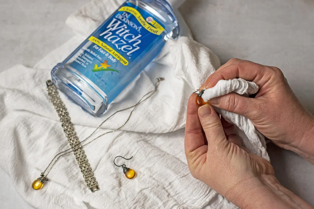 Hands are cleaning an earring with a white cloth. A bottle of witch hazel is laying in the background next to the other earring, a necklace and a bracelet. 