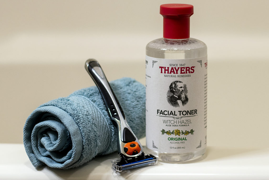 A rolled up washcloth, a men's razor and a bottle of Thayer's witch hazel sit on the edge of a bathtub.