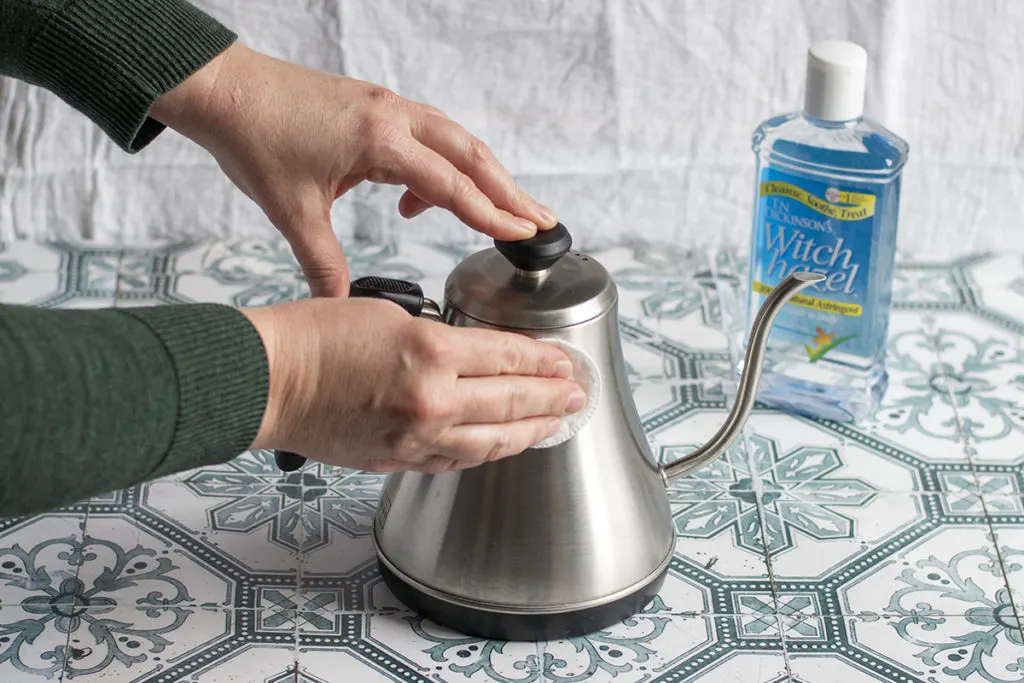 Hands are shown using a cotton round and witch hazel to clean a stainless steel electric kettle.