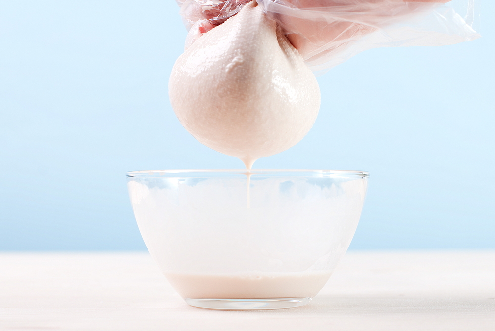 A gloved hand squeezes a nut milk bag over a bowl making almond milk. 