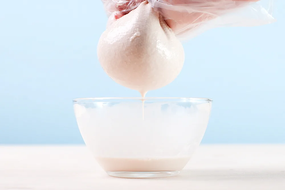 A gloved hand squeezes a nut milk bag over a bowl making almond milk. 