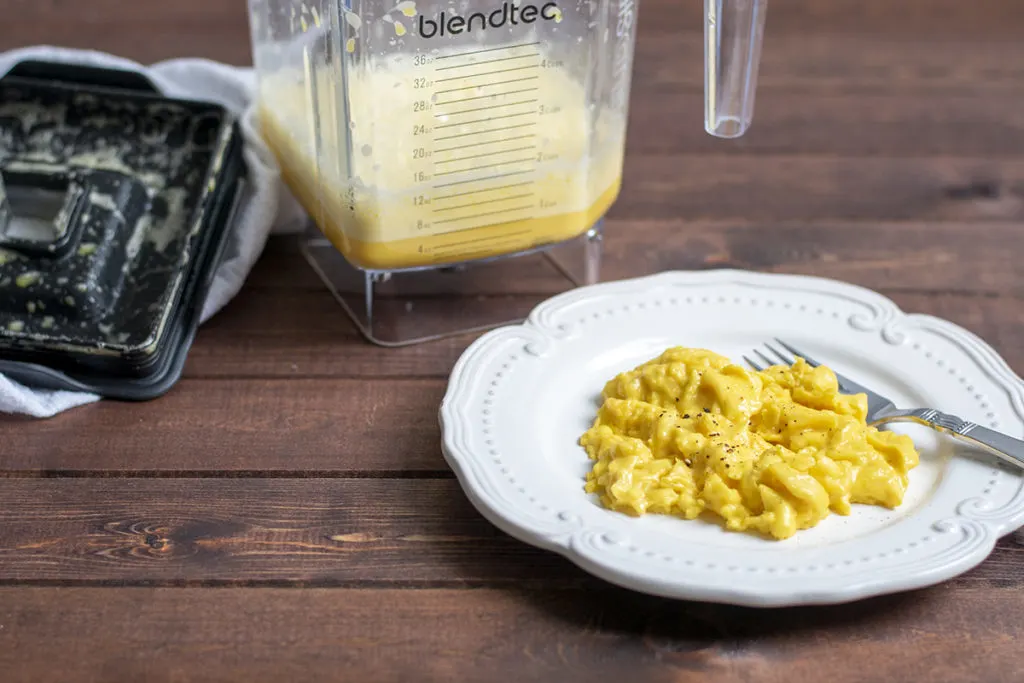 A plate of scrambled eggs with a fork. There is a blender jar behind them with raw eggs in the jar.