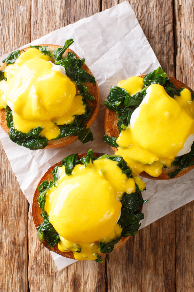 Three eggs benedict on parchment paper on a wood background.