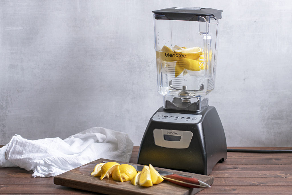 A blender with lemons ice and water in it. There are fresh cut lemons on a cutting board next to a knife in front of the blender. 
