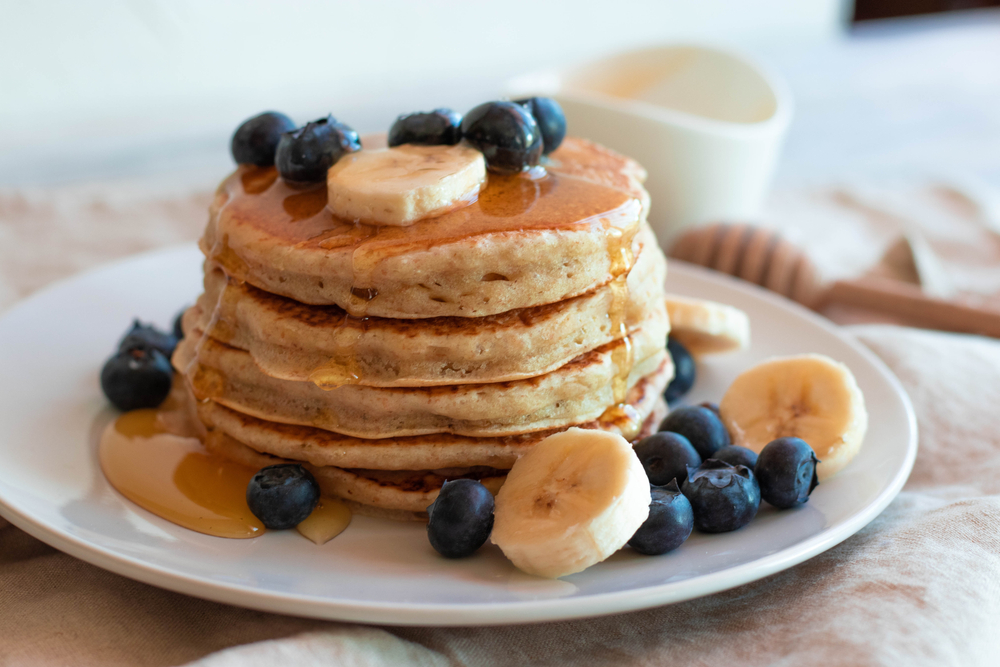 A stack of pancakes decoratively arranged on a plate. The pancakes are drizzled with maple syrup, bananas and blueberries.