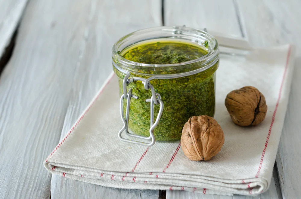 A jar of pesto sitting on a folded tea towel next to two whole walnuts.