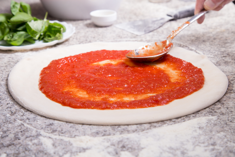 A spoon is spreading pizza sauce on pizza dough.