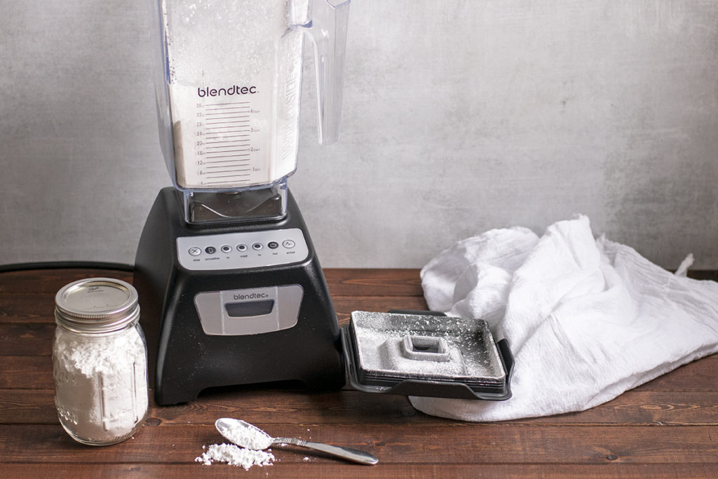 A blender with powdered sugar in the jar. A mason jar with powdered sugar in it and a spoonful of powdered sugar on a wooden top, a white tea towel is laying in the background.