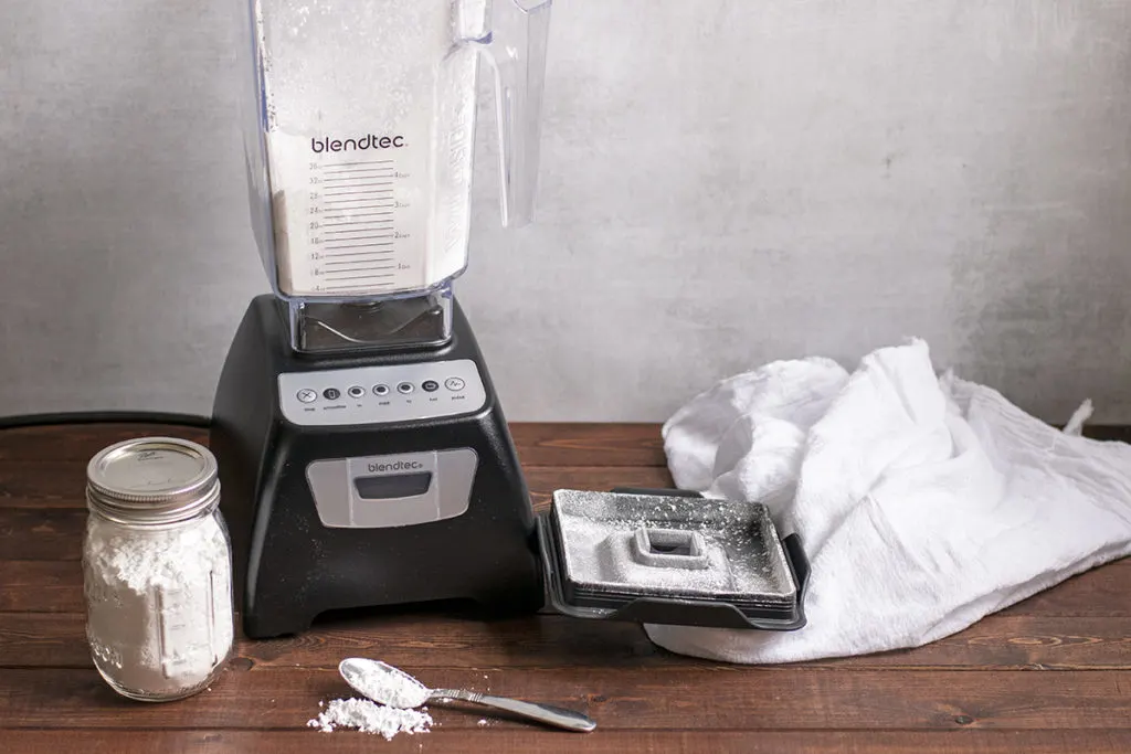 A blender with powdered sugar in the jar. A mason jar with powdered sugar in it and a spoonful of powdered sugar on a wooden top, a white tea towel is laying in the background.
