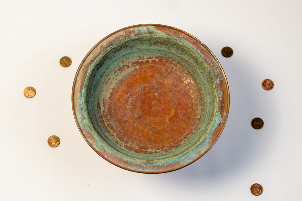A copper dish is covered in green oxidation. There are pennies scattered around the outside of the dish. 