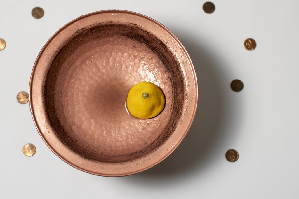 Half of a lemon is sitting in a copper dish, there are pennies scattered around the outside of the dish.
