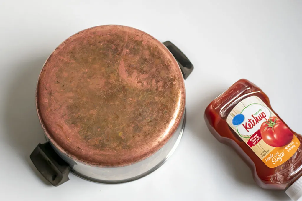 A copper bottomed pot is upside down with a ketchup bottle next to it. 
