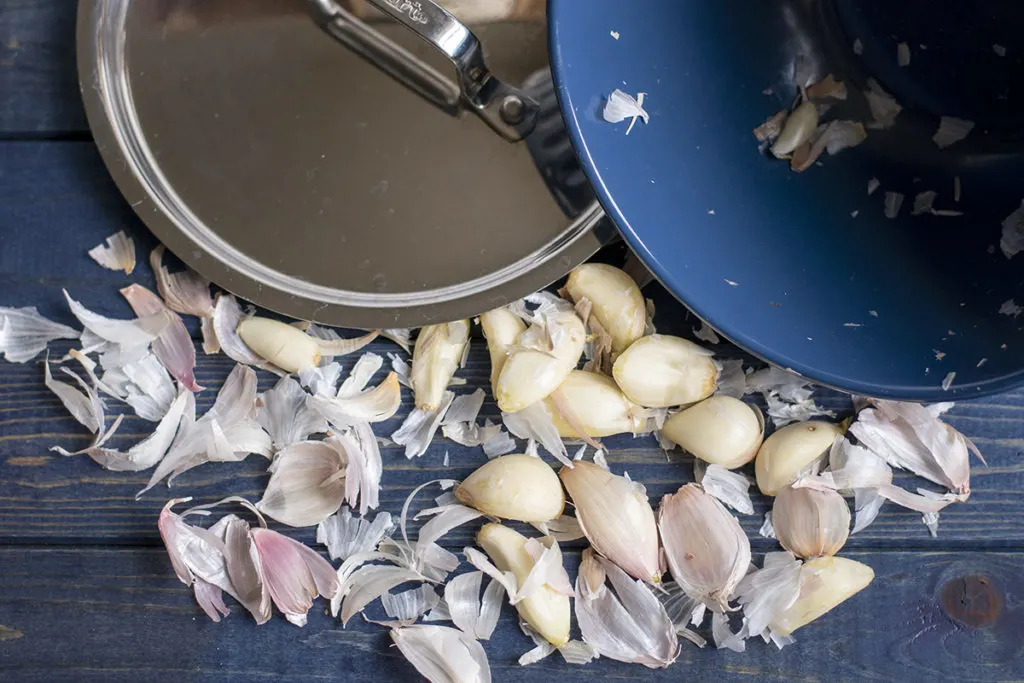 a metal pot lid, the empty blue bowl and peeled garlic cloves and their skins are decoratively arranged on a blue wood tabletop.