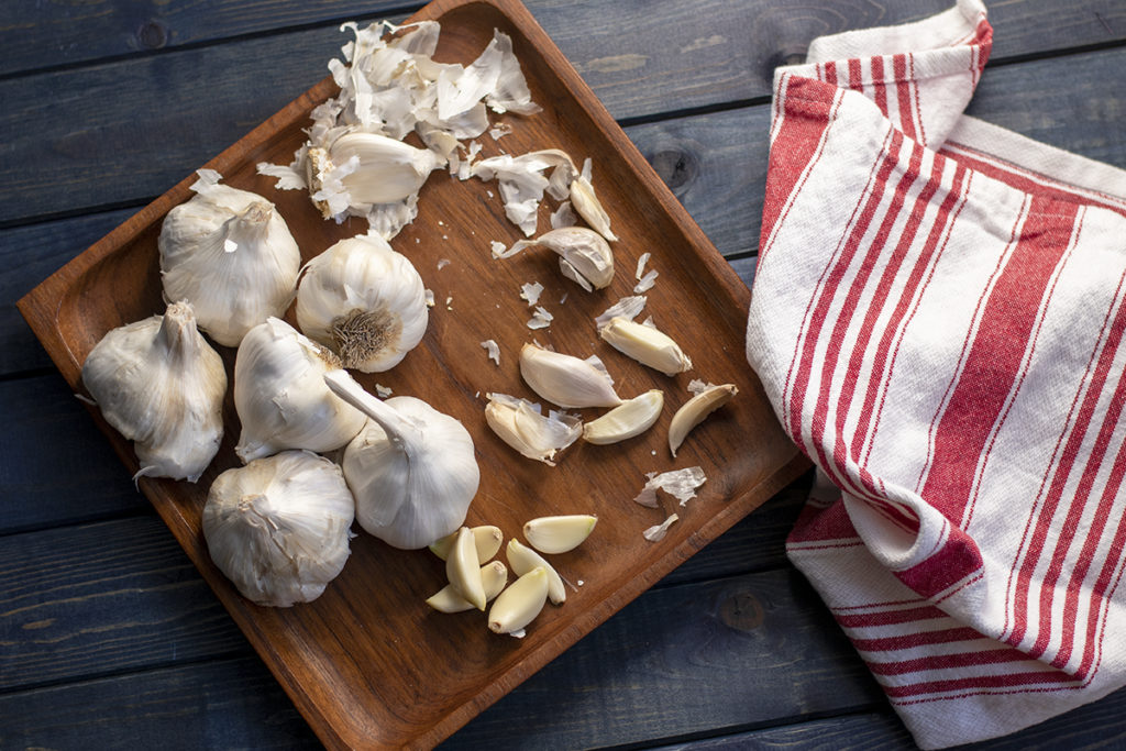 Garlic heads and cloves, peeled and unpeeled on a teak platter next to a red and white tea towel.