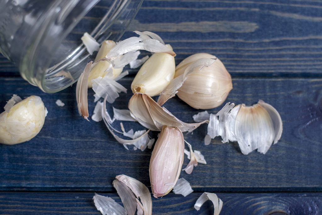 a clear jar emptied of the peeled garlic cloves and skins on a blue wood background.