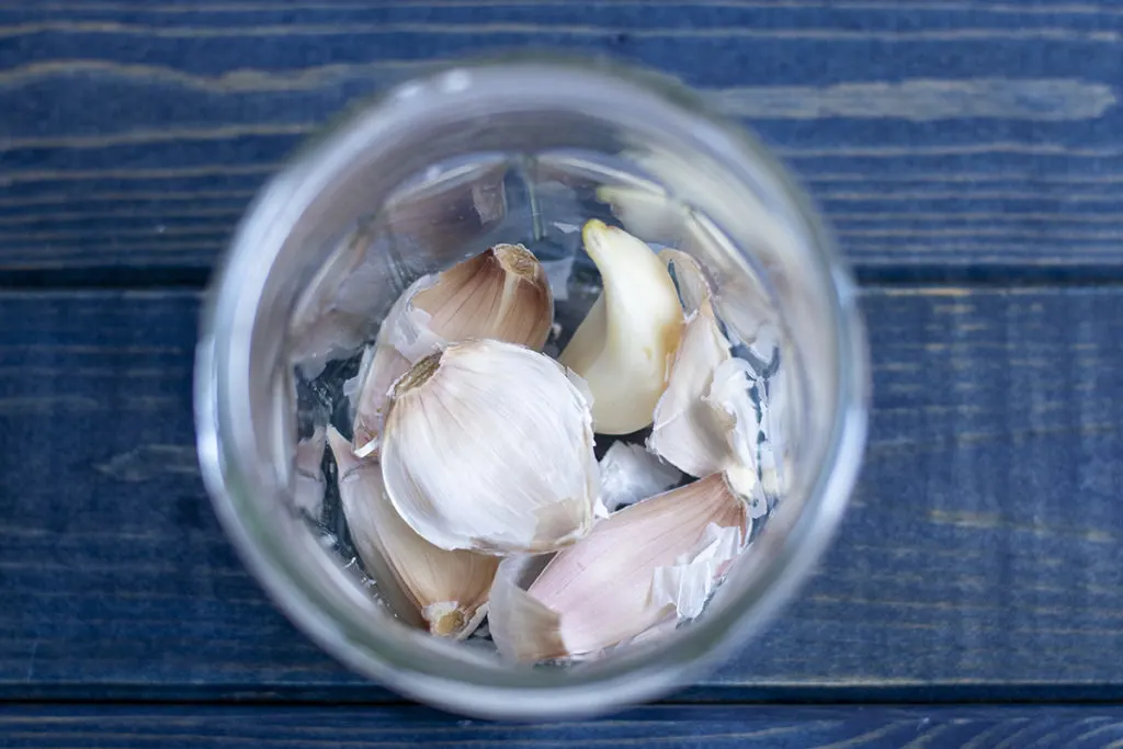 Half-peeled garlic cloves in a clear jar on a blue wood background.
