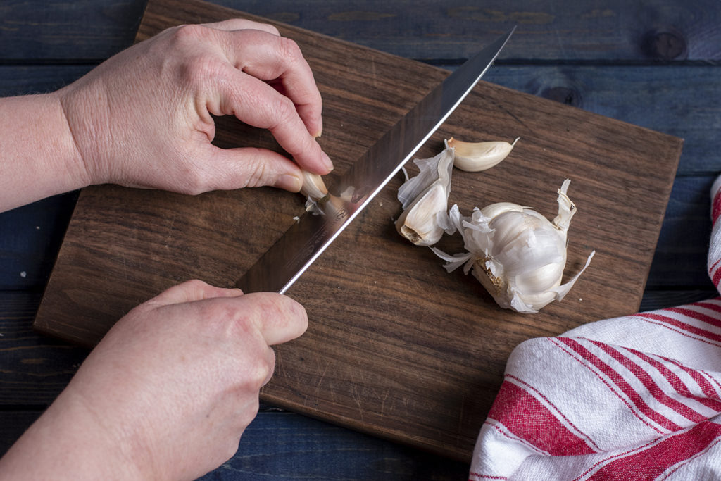 I am using a chef knife to cut the root tip off of a clove of garlic on a cutting board. The rest of the bulb of garlic is setting next to my hands. 