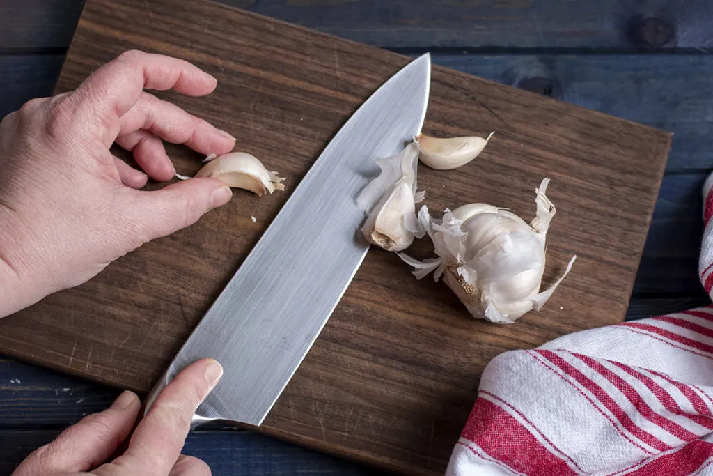 I am positioning a garlic clove on the cutting board with one hand and holding a chef knife with the other.