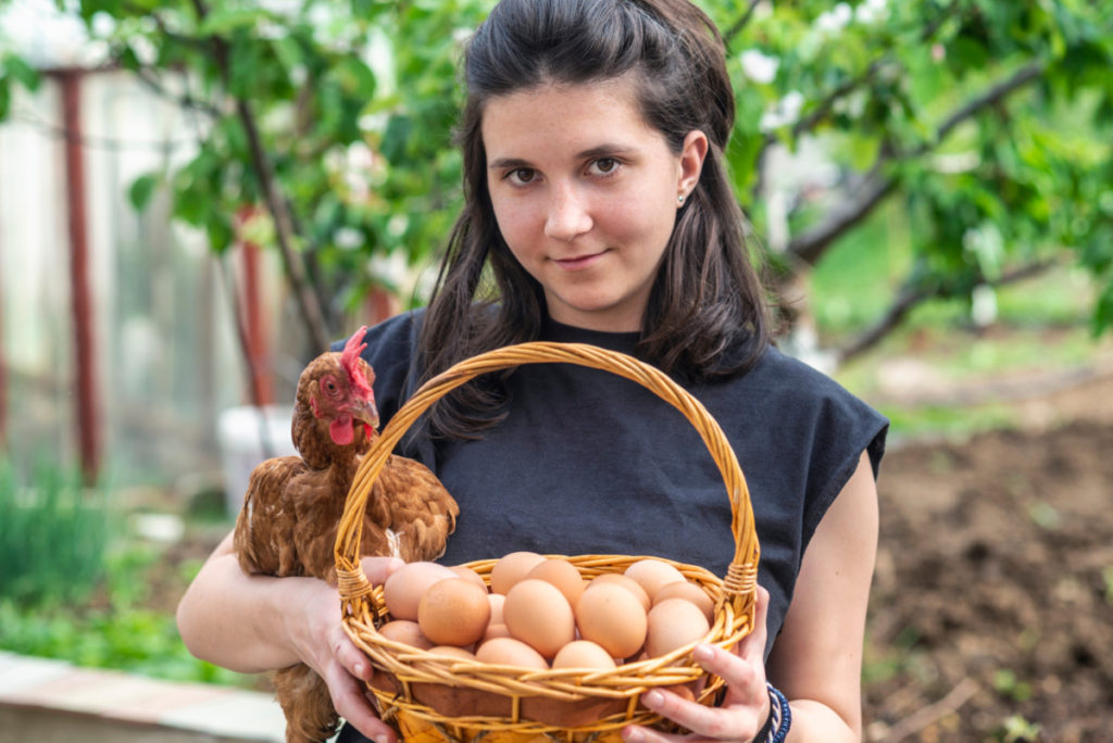 A young woman holds a chicken under her arm and a basket of eggs. She is smiling at the camera.