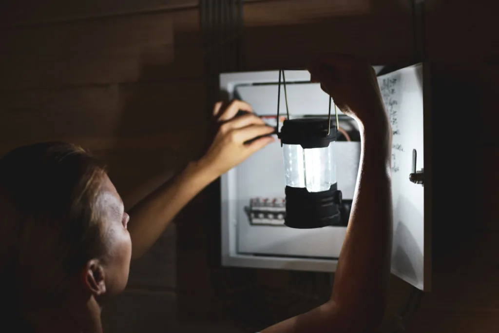 A woman holding a battery powered lantern up to a breaker box in the dark. 