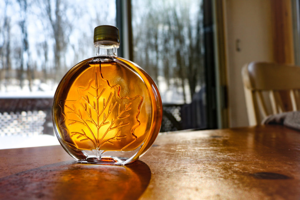 A bottle of maple syrup catches the sun in front of a window that looks out on a snowy day.
