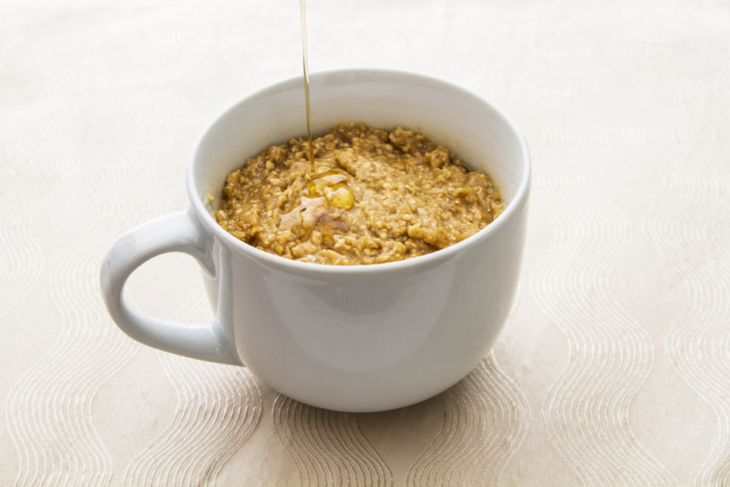 A mug with oatmeal in it, maple syrup is being drizzled over top.