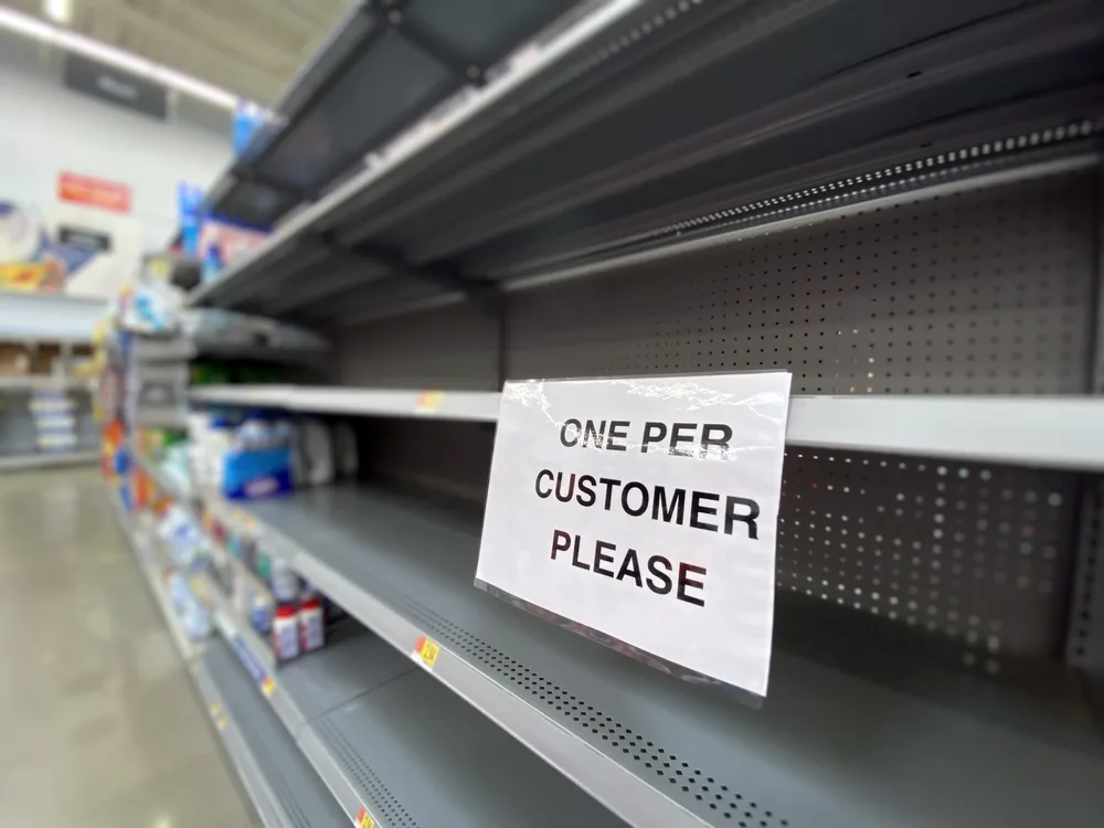 Bare grocery store shelves are shown with a sign that read, 'One Per Customer Please'