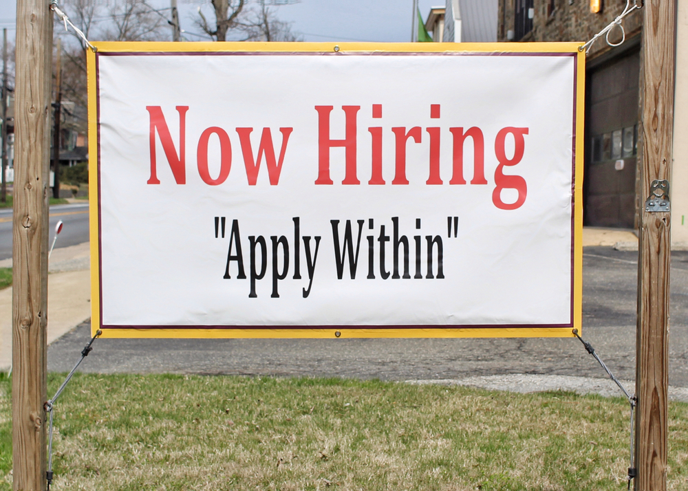 A sign hanging outdoors reads, "Now Hiring Apply Within"