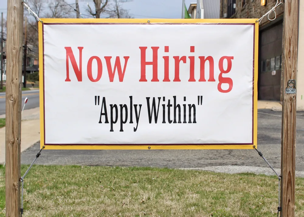 A sign hanging outdoors reads, "Now Hiring Apply Within"